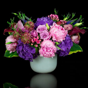 Elegant floral arrangement featuring purple hydrangeas, pink peonies, and roses in a white vase against a black background.