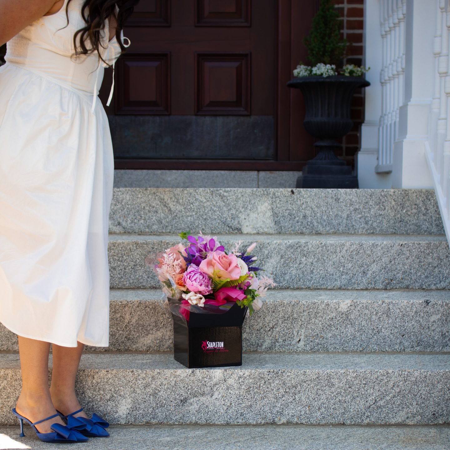 Flower delivery by Boston florist Stapleton Floral, featuring a vibrant bouquet on a doorstep with a woman in a white dress and blue heels nearby.