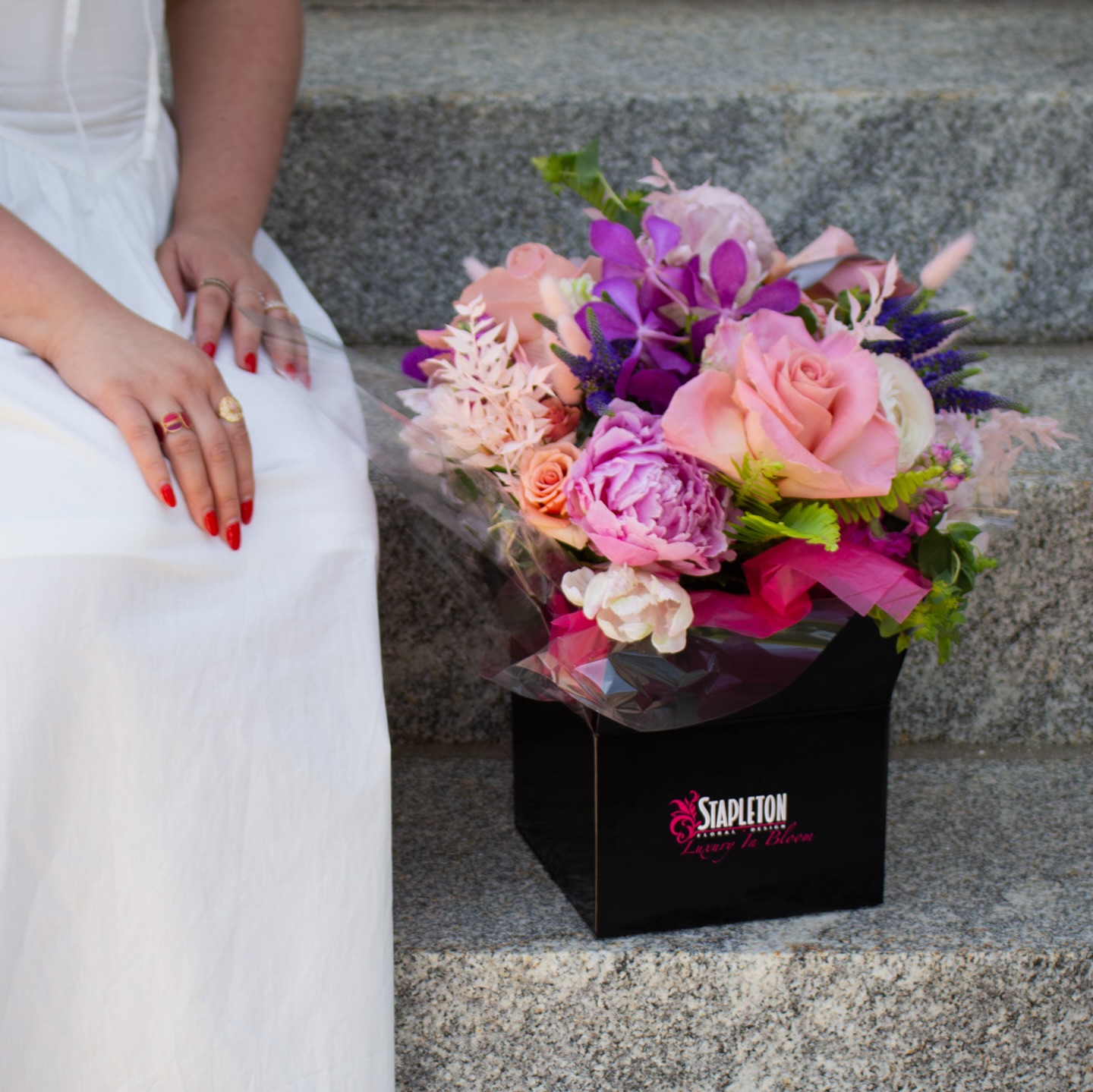 Close-up of a vibrant floral arrangement from Boston florist Stapleton Floral in a black box, placed on granite steps next to a woman in a white dress with red nails.