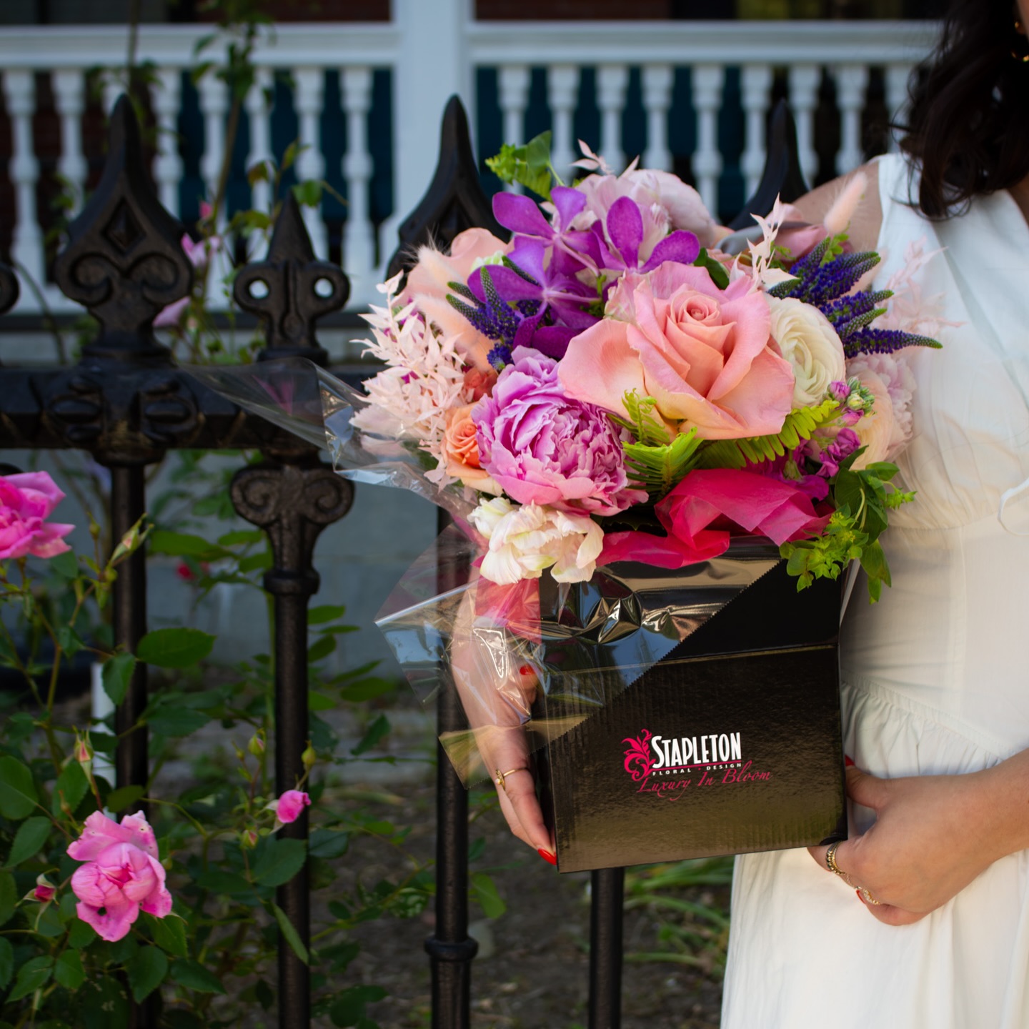 Close-up of a woman in a white dress holding a black Stapleton Floral arrangement box filled with vibrant roses, peonies, and orchids in pink and purple hues, with a garden fence behind.