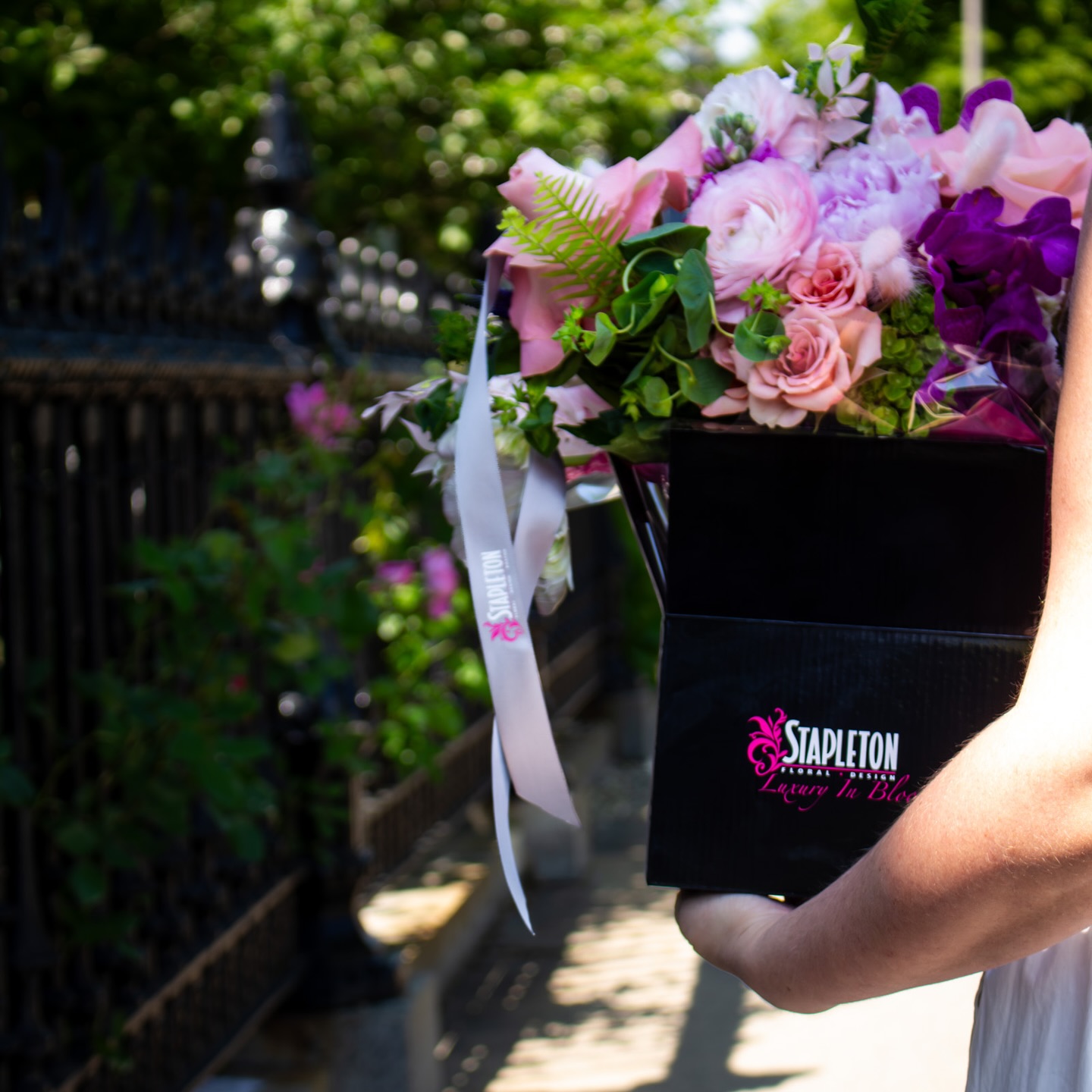 A person holding a luxury black Stapleton Floral flower box filled with pink and purple seasonal blooms, walking along a sunny Boston street with wrought iron fencing in the background.
