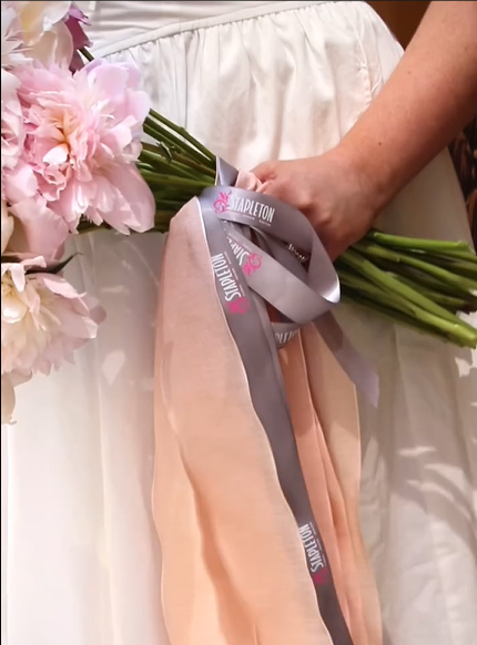Woman holding a bouquet of pink flowers wrapped in Stapleton Floral ribbon with soft peach and gray fabric, wearing a white dress.