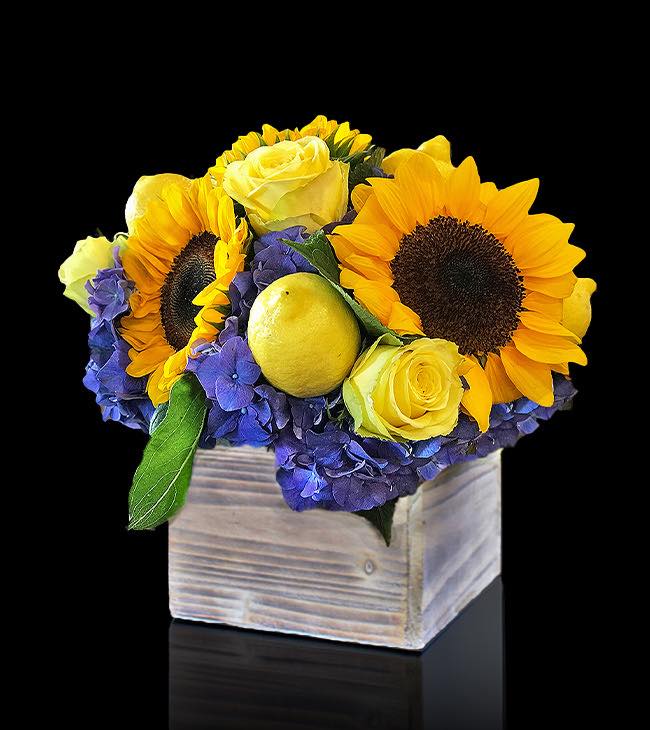 A vibrant Lemon Drop flower arrangement featuring sunflowers, yellow roses, purple hydrangeas, and real lemons in a rustic wooden box.