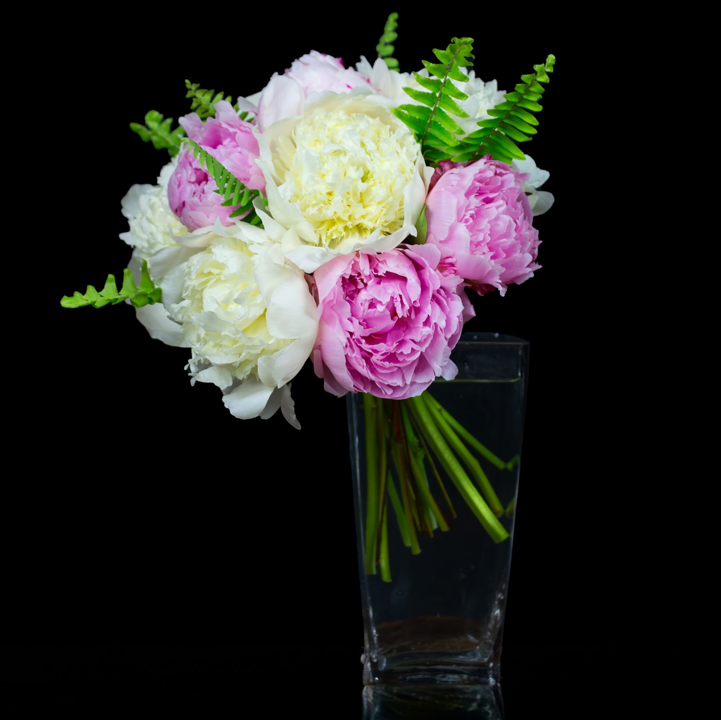 Lush bouquet of white and pink peonies with fern accents arranged in a clear hexagonal vase against a black background.