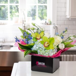 Stapleton Floral hand-delivered arrangement featuring orchids, hydrangeas, and roses in branded packaging on a bright kitchen counter.