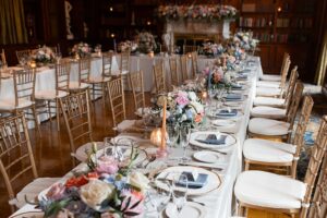 Long wedding reception table with gold chairs, pastel floral centerpieces, and elegant place settings in a warm, intimate venue.