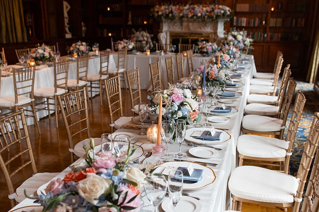 Long wedding reception table with gold chairs, pastel floral centerpieces, and elegant place settings in a warm, intimate venue.