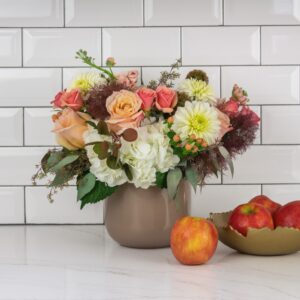 A beautiful flower arrangement featuring roses, dahlias, hydrangeas, and eucalyptus, set against a white tiled wall with apples in the foreground. Perfect for brightening any space in Boston.