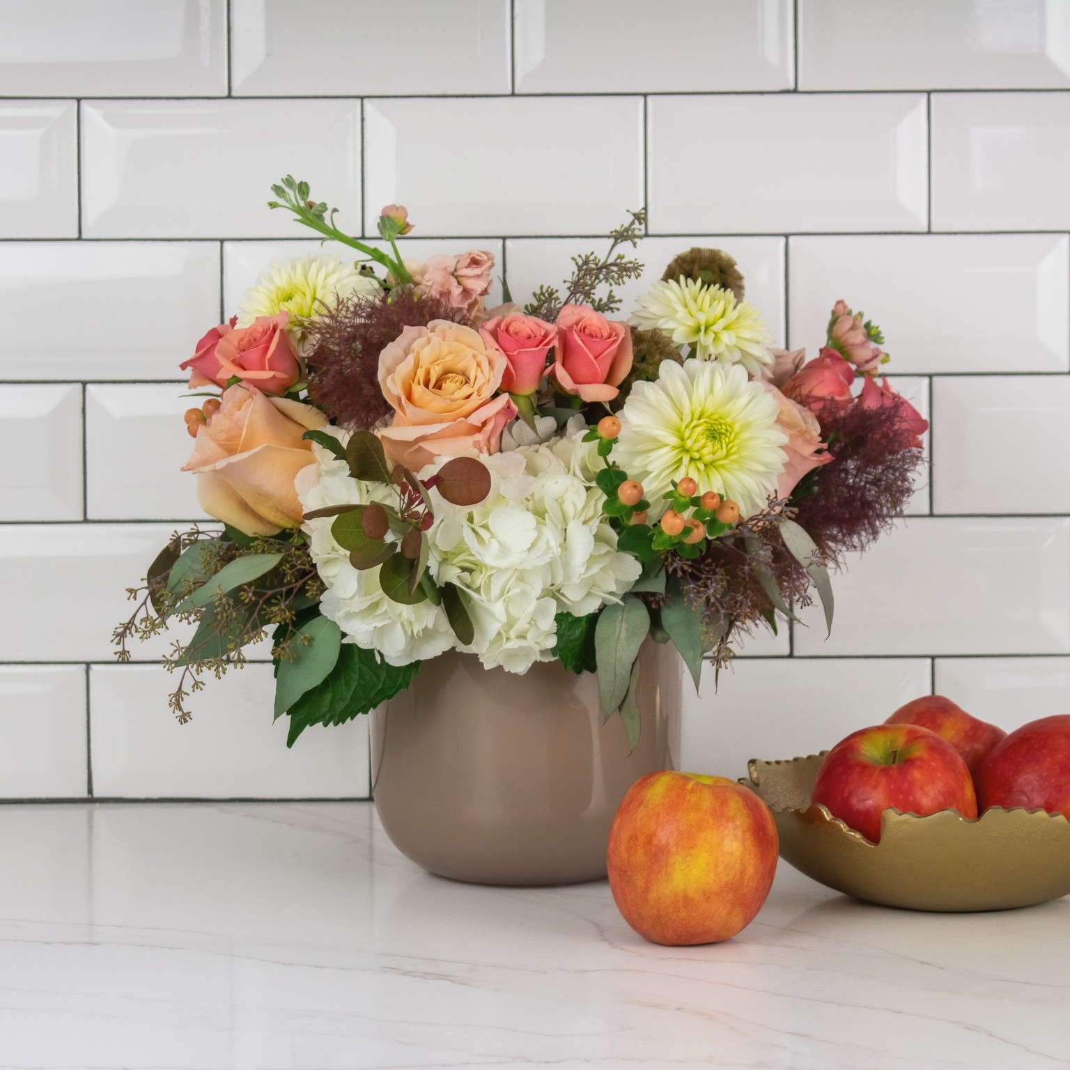 A beautiful flower arrangement featuring roses, dahlias, hydrangeas, and eucalyptus, set against a white tiled wall with apples in the foreground. Perfect for brightening any space in Boston.