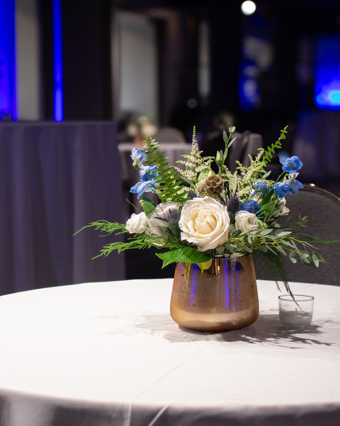Close-up of a blue and white floral arrangement in a gold vase on a white tablecloth, styled for a cocktail reception.
