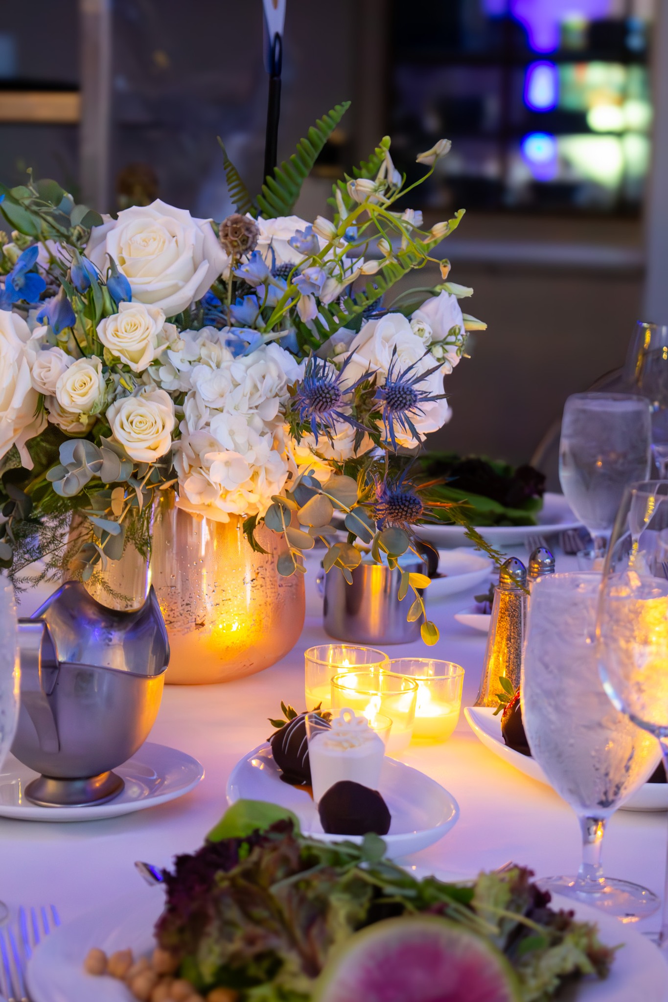 Blue and white floral centerpiece with roses and thistles in a glowing vase, surrounded by candles and dinner settings at an event.