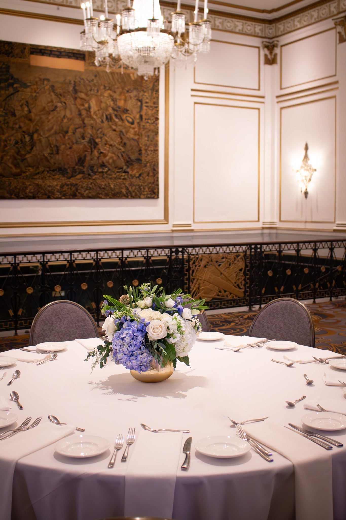 Event floral arrangement of blue hydrangeas, white roses, and greenery in a gold vase, styled for a banquet setting.