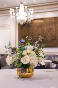 Elegant event floral centerpiece with white roses, blue accents, and greenery in a gold vase, displayed under a chandelier.