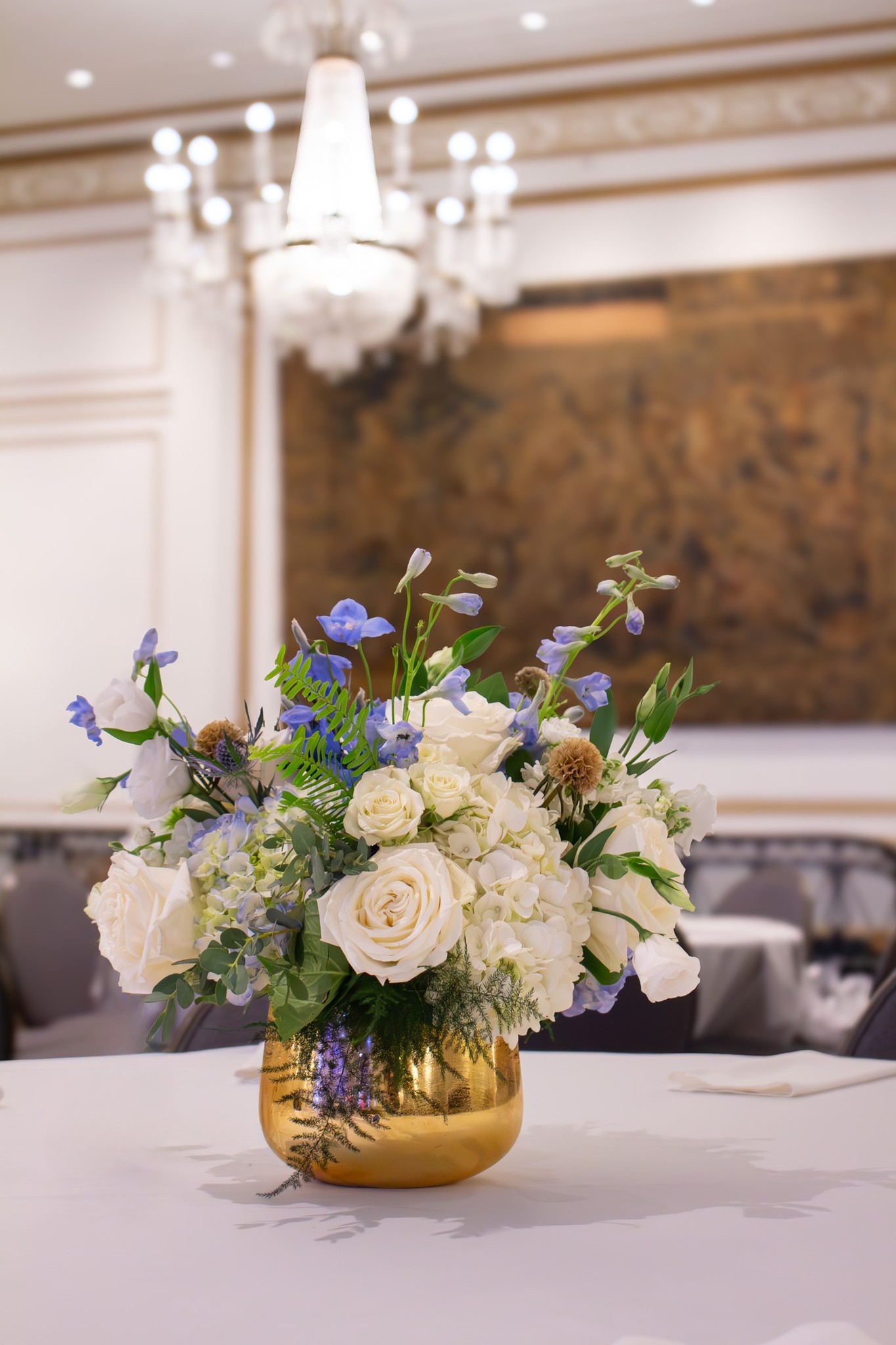 Elegant event floral centerpiece with white roses, blue accents, and greenery in a gold vase, displayed under a chandelier.