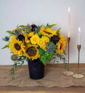 Golden Twilight floral arrangement featuring bright yellow sunflowers, roses, and autumn textures in a black vase beside two gold candlesticks on a burlap-covered table.