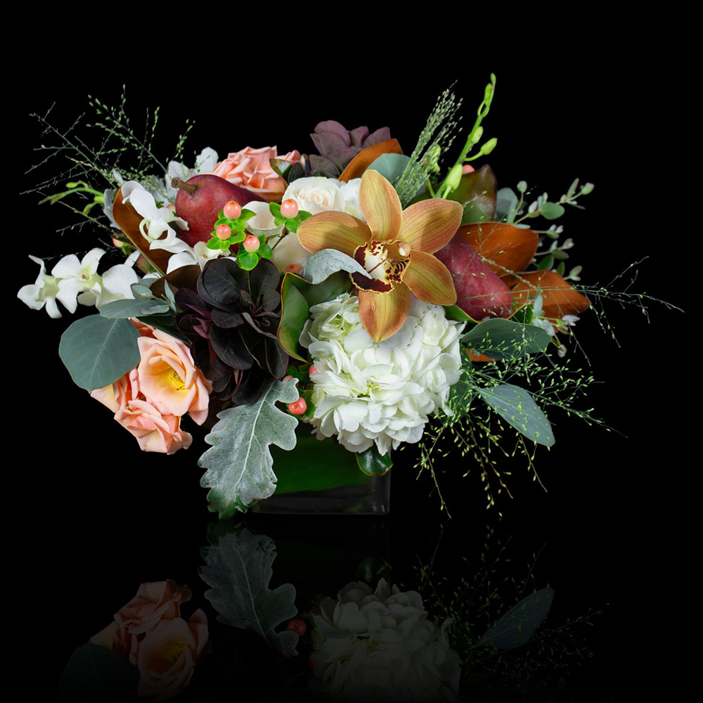 Side view of the Harvest Hearth Thanksgiving floral centerpiece arranged with roses, hydrangea, orchids, pears, and fall foliage in a modern vase on a black background.