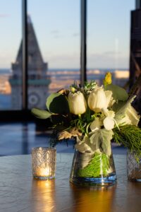 Winter white floral arrangement with evergreen foliage and candlelight, styled on a tabletop with a city skyline visible through large windows.